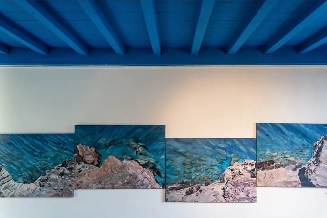 Het meerdelige schilderij Itanos aan een witte tentoonstellingswand onder een blauw plafond.